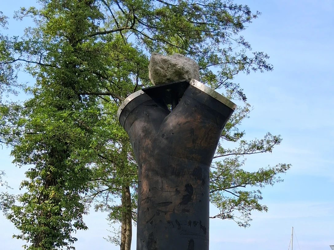 Luft-Skulptur-Skulpturenpromenade-Steinhude3 Skulptur aus dunklem Metall auf grünem Gras vor Bäumen und See. Im Hintergrund liegt ein Bootssteg.