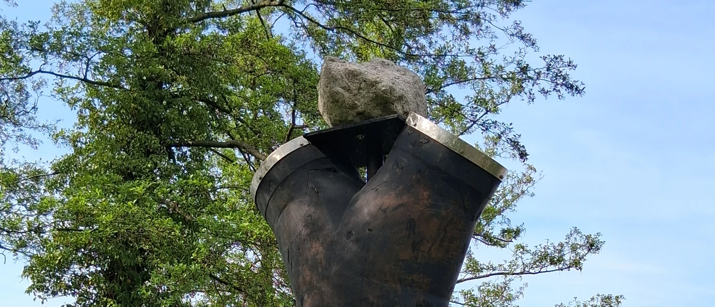 Luft-Skulptur-Skulpturenpromenade-Steinhude3 Skulptur aus dunklem Metall auf grünem Gras vor Bäumen und See. Im Hintergrund liegt ein Bootssteg.