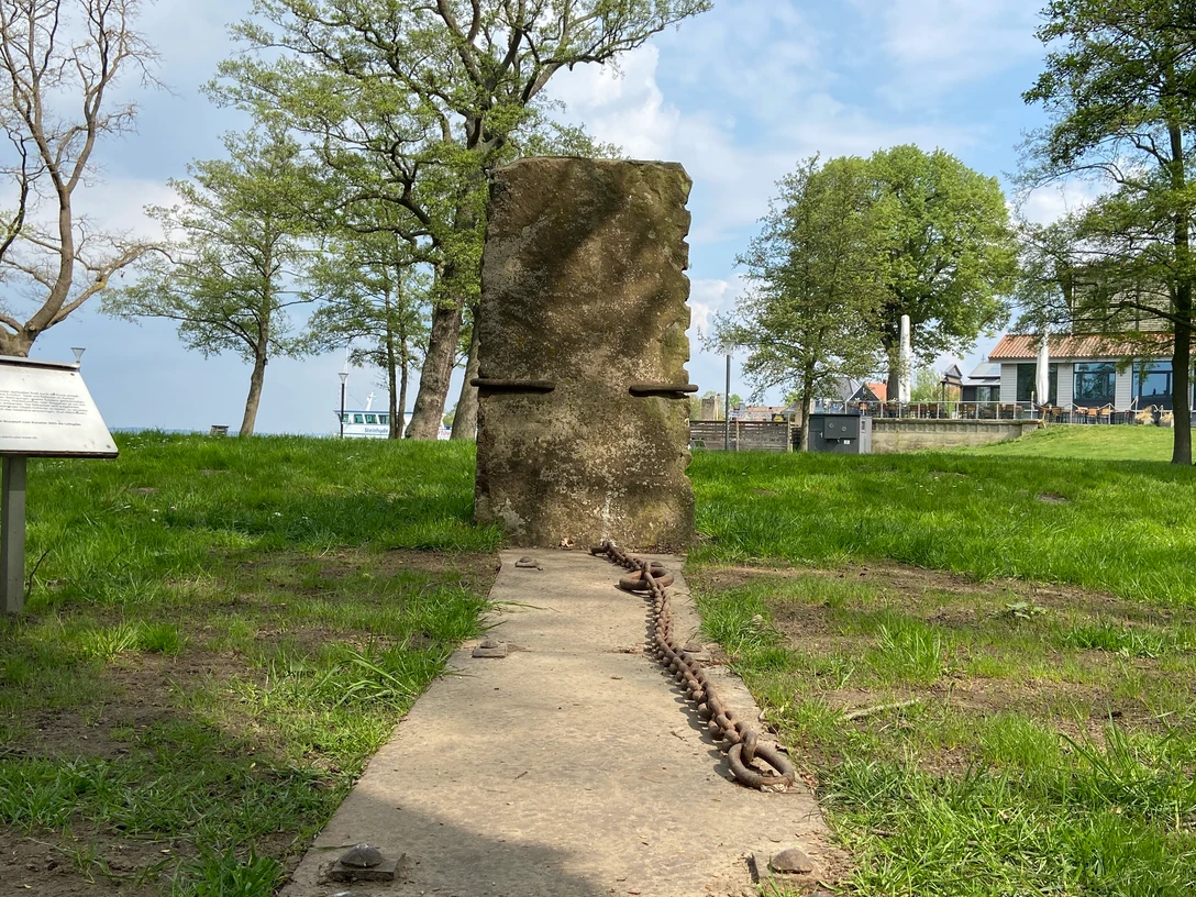 Hinführung-Skulptur-Skulpturenpromenade-Steinhude3 Steinhude Skulpturenpromenade: Aufrecht stehende Steinplatte mit Ketten auf einer Wiese am Seeufer.Steinhude sculpture promenade: Upright stone slab with chains on a meadow on the lakeshore.Steinhudes skulpturpromenade: Opretstående stenplade med kæder på en eng ved søbredden.Steinhude sculptuur promenade: Rechtopstaande stenen plaat met kettingen op een weiland aan de oever van het meer.