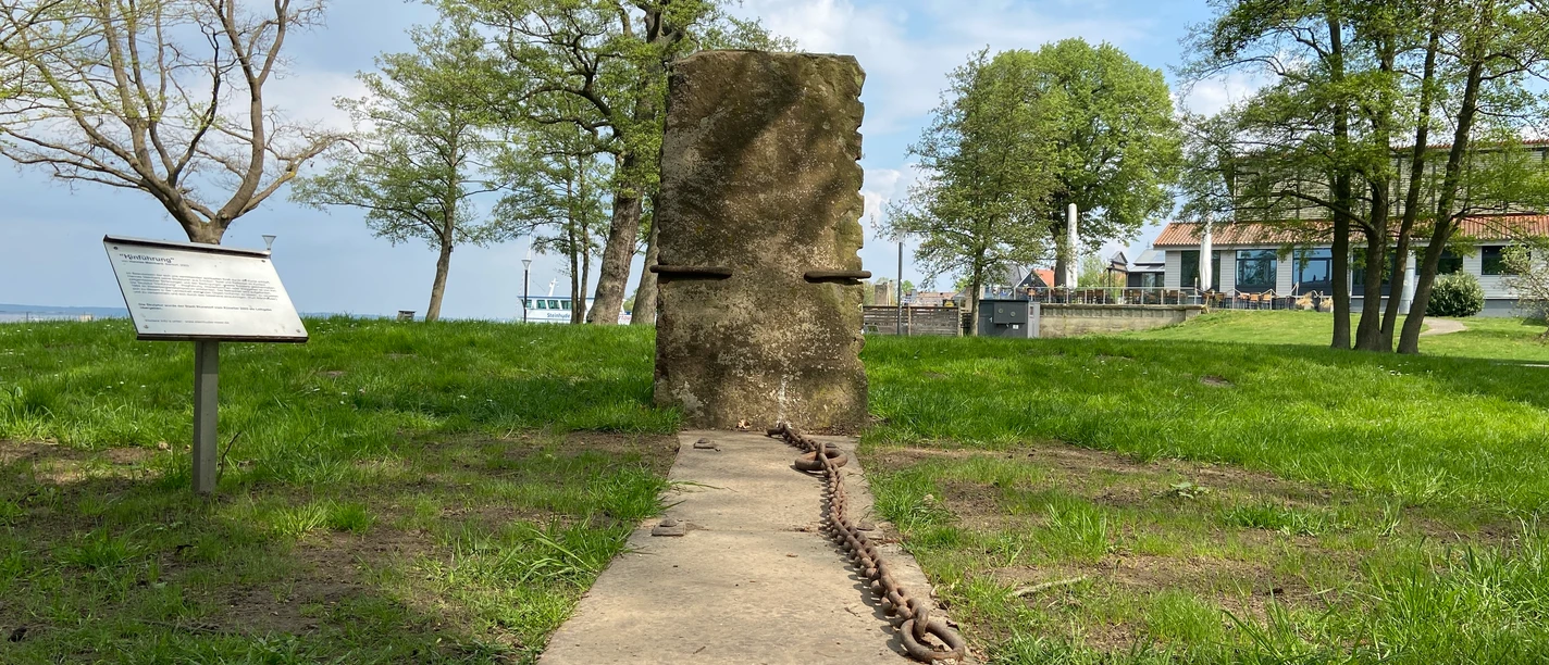 Hinführung-Skulptur-Skulpturenpromenade-Steinhude3 Steinhude Skulpturenpromenade: Aufrecht stehende Steinplatte mit Ketten auf einer Wiese am Seeufer.