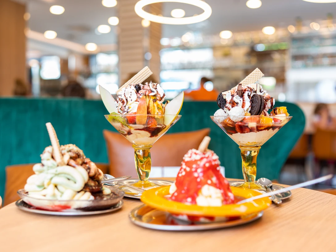 Eiscafé Dolomiti Drei kunstvoll angerichtete Eisbecher mit verschiedenen Früchten und Toppings auf einem Holztisch.