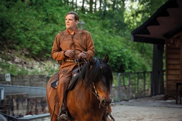 A man in historical western clothing rides a brown horse in front of a green backdrop.
