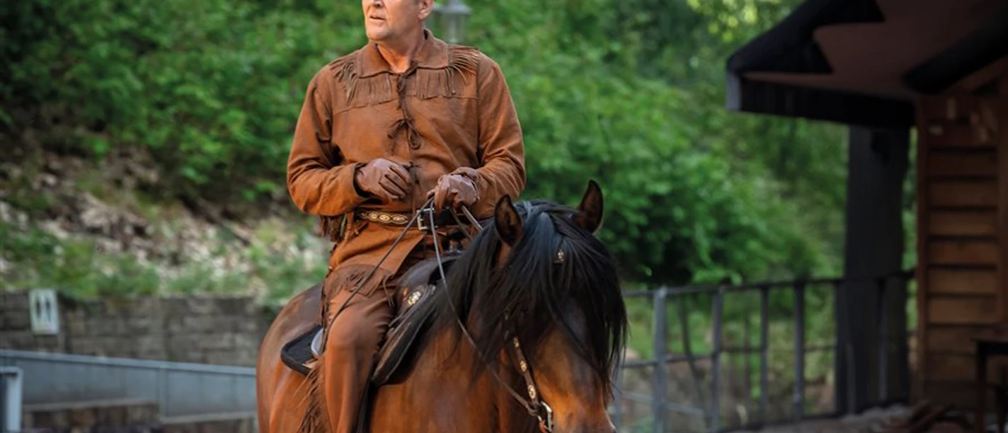 Shatterhand_0_Landesbuehnen-Sachsen_900x600.jpg Ein Mann in historischer Westernkleidung reitet konzentriert auf einem braunen Pferd vor grüner Kulisse.
