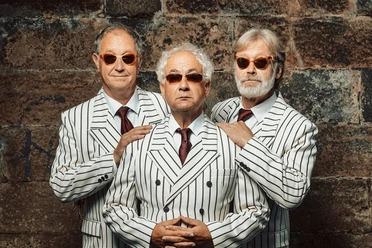 Drie oudere mannen in witte pakken en rode dassen staan voor een stenen muur.