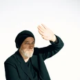 straeter_torsten_marvin_ruppert_.png Älterer Mann mit grauem Bart und schwarzer Kleidung hebt die Hand in hellem, minimalistischen Umfeld.