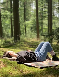 Waldbaden3.jpeg Frau entspannt auf Picknickdecke in einem lichtdurchfluteten Wald, umgeben von hohen Bäumen.