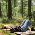 Waldbaden3.jpeg Frau entspannt auf Picknickdecke in einem lichtdurchfluteten Wald, umgeben von hohen Bäumen.