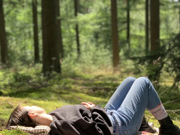 Waldbaden3.jpeg Frau entspannt auf Picknickdecke in einem lichtdurchfluteten Wald, umgeben von hohen Bäumen.