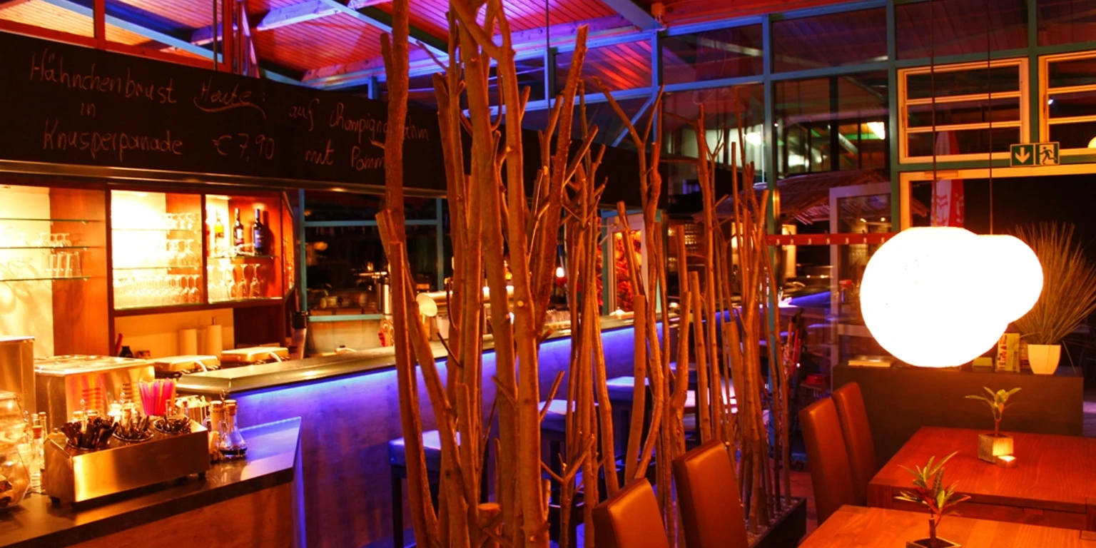 Raumfoto_Tresen.JPG Ein stilvoll beleuchteter Barbereich mit farbigem Licht, modernen Holzmöbeln und dekorativen Zweigen.