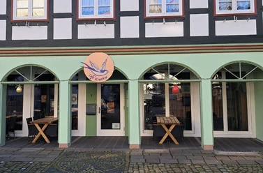1b4c911c-d858-4fb6-b3b0-0cdaa397952b.jpg Die Fassade eines Fachwerkhauses mit vier grünen Rundbogenfenstern und einem leuchtenden Café-Schild.