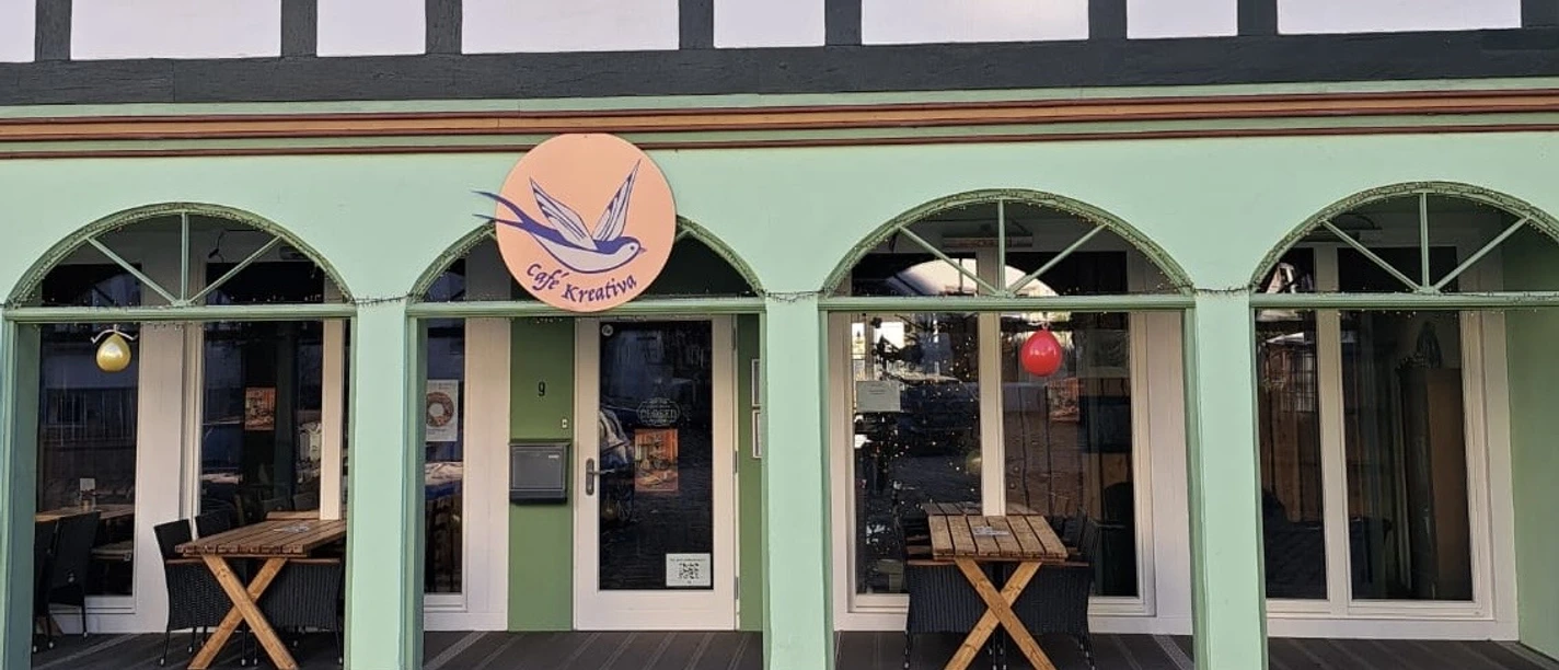 1b4c911c-d858-4fb6-b3b0-0cdaa397952b.jpg Die Fassade eines Fachwerkhauses mit vier grünen Rundbogenfenstern und einem leuchtenden Café-Schild.