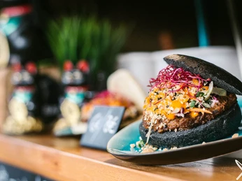 Street Food Festival Köln Ein saftiger Burger mit schwarzem Brötchen, frischen Beilagen und leuchtendem Gemüse wird appetitlich auf einem Teller präsentiert.A juicy burger with a black bun, fresh garnishes and bright vegetables is appetizingly presented on a plate.