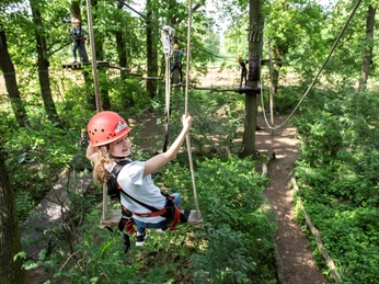 Eine junge Frau schwingt sich in einem Kletterpark zwischen Bäumen, gesichert mit Helm und Gurt.A young woman swings between trees in a climbing park, secured with a helmet and harness.En ung kvinde svinger sig mellem træerne i en klatrepark, sikret med hjelm og sele.Een jonge vrouw schommelt tussen bomen in een klimpark, gezekerd met een helm en een harnas.