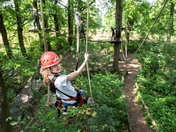 A young woman swings between trees in a climbing park, secured with a helmet and harness.