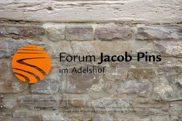 forum-jacobpins-schild Schild aus Metall mit orangefarbenem Symbol und schwarzer Schrift auf aus Stein gemauerter Wand.