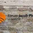 forum-jacobpins-schild Schild aus Metall mit orangefarbenem Symbol und schwarzer Schrift auf aus Stein gemauerter Wand.