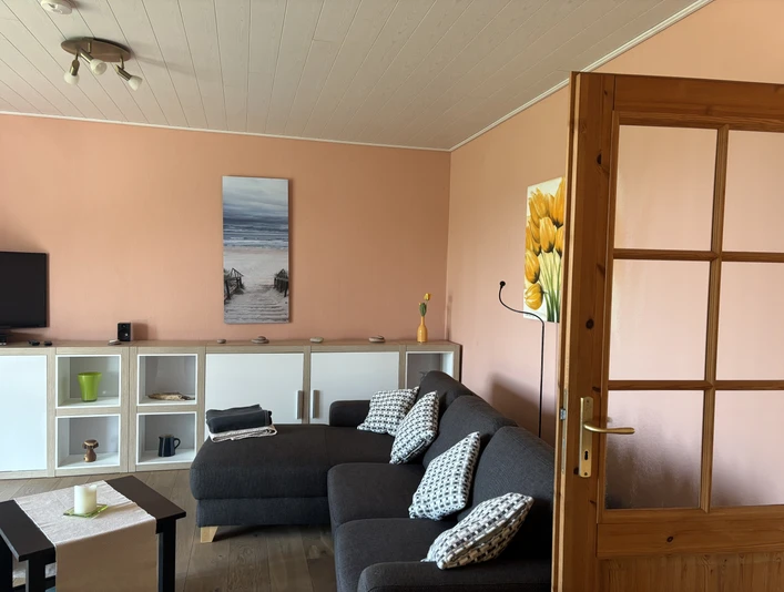 Ferienhaus im Öspergarten Wohnzimmer einer Ferienwohnung mit grauem Ecksofa, Pastellwänden und weißen Regalen.