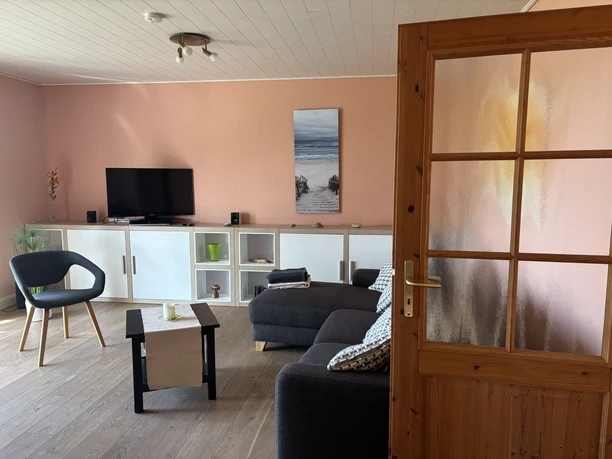 Wohnzimmer mit hellen Holzfußböden, rosa Wänden und einer modernen, grauen Eckcouch.