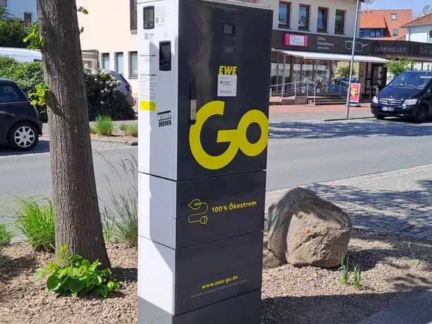 EWE-Go-Ladesäule an einer Straße, neben Baum und Gehweg.