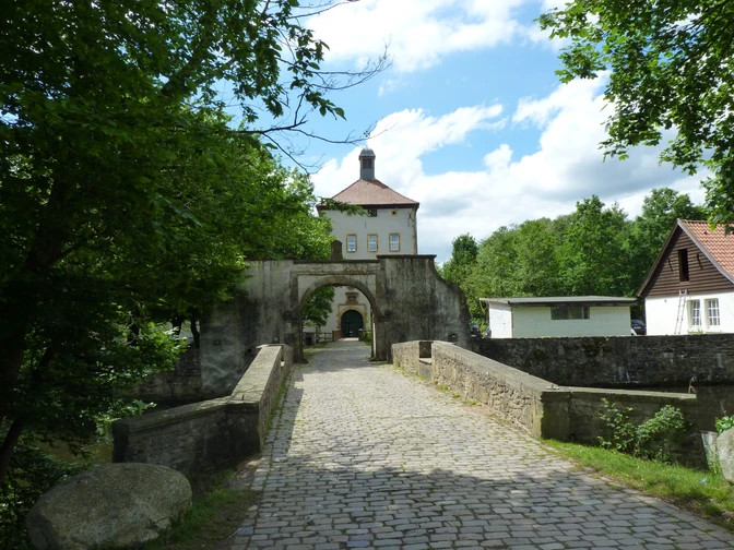 Historisches Gut mit Brücke und Fachwerkgebäude umgeben von üppigem Grün unter blauem Himmel.