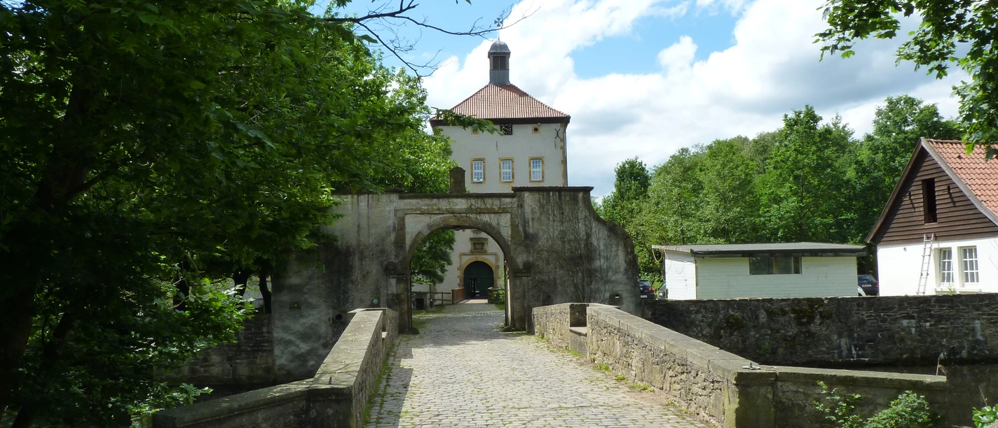 POI-8-e_Gut-Bustedt.JPG Historisches Gut mit Brücke und Fachwerkgebäude umgeben von üppigem Grün unter blauem Himmel.