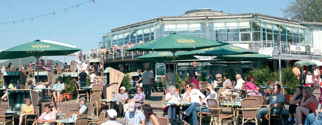 Eine belebte Café-Terrasse mit vielen Menschen, die an Tischen unter grünen Sonnenschirmen sitzen.