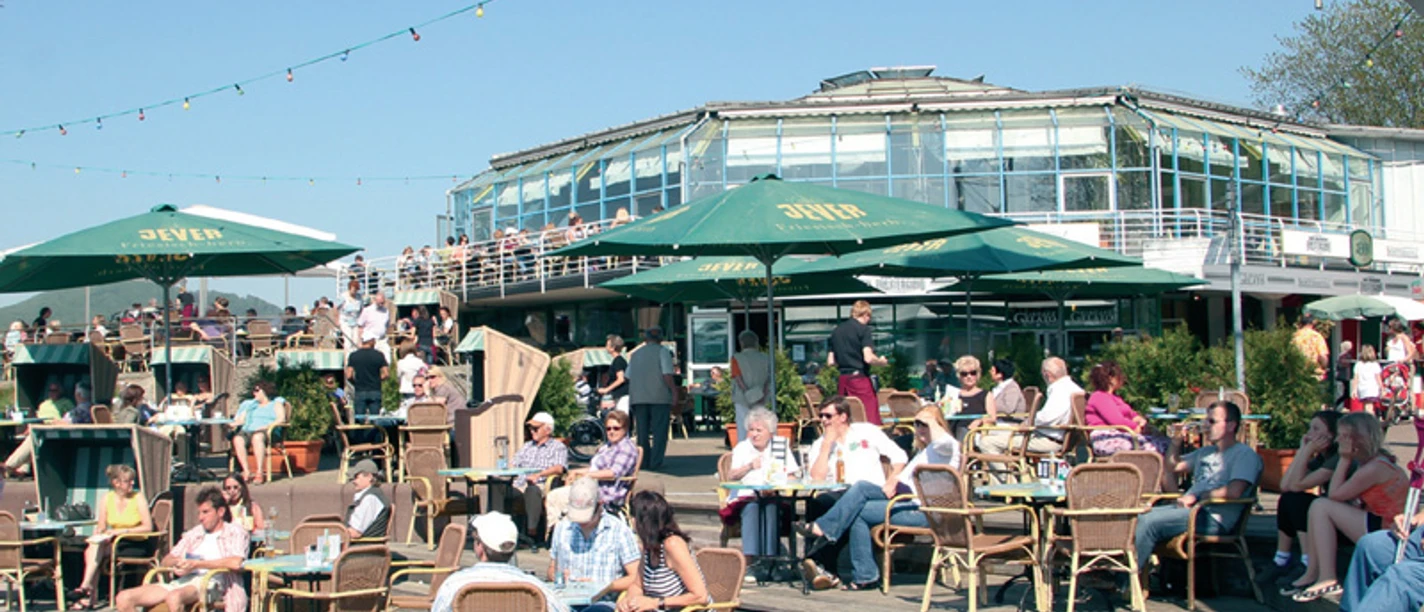 Eine belebte Café-Terrasse mit vielen Menschen, die an Tischen unter grünen Sonnenschirmen sitzen.