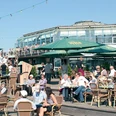 Café Breitengrad.jpg Eine belebte Café-Terrasse mit vielen Menschen, die an Tischen unter grünen Sonnenschirmen sitzen.