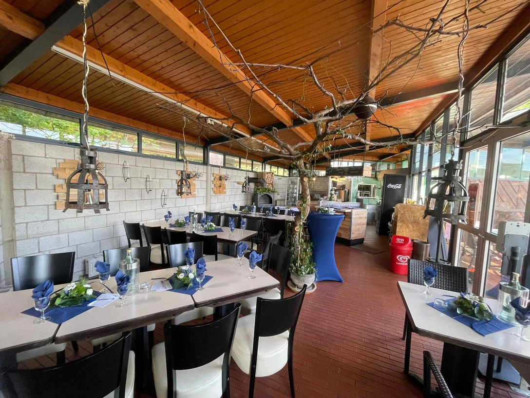 IMG-20220917-WA0007.jpg Ein luftiger Raum mit Holzdecke, elegant gedeckten Tischen und einem rustikalen Ofen als Blickpunkt.