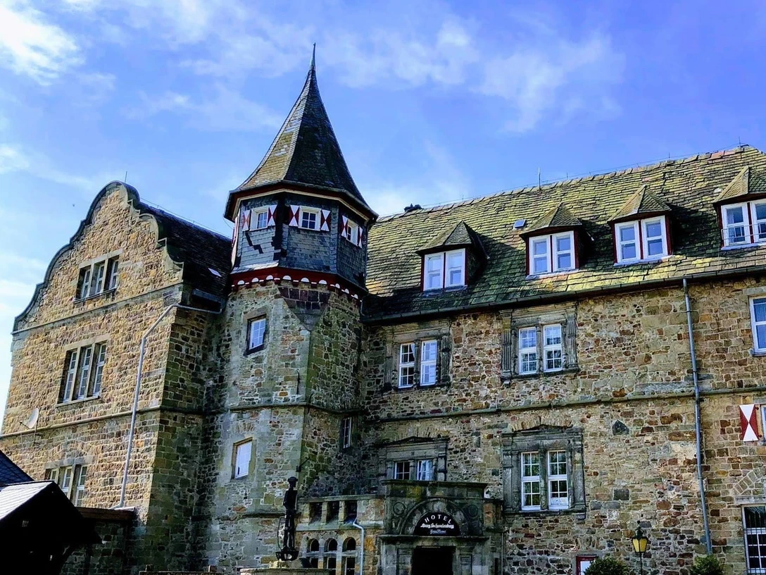 IMG_2105.jpg Historisches Steingebäude mit Turm und Dachgauben, umgeben von blühenden Pflanzen, unter blauem Himmel.