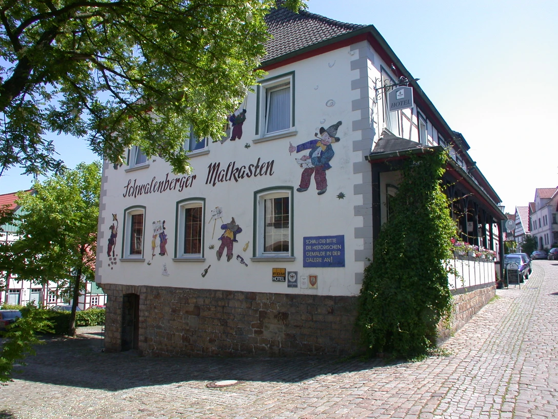 Fachwerkgebäude mit Wandbemalung und Figurendekor. Kopfsteinpflaster und sonnige Umgebung ergänzen das Bild.