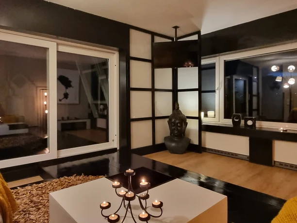 Wohnzimmer.jpg Modernes Wohnzimmer mit großen Glasfenstern, Kerzenlicht und Buddha-Statue für entspannte Atmosphäre.