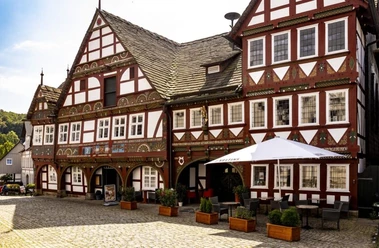 exterieurCL9A9992-2000-1-e1724078820263-768x502.jpg Historisches Fachwerkhaus mit verzierten Fassaden, Arkadengang und Außensitzbereich auf gepflastertem Platz.