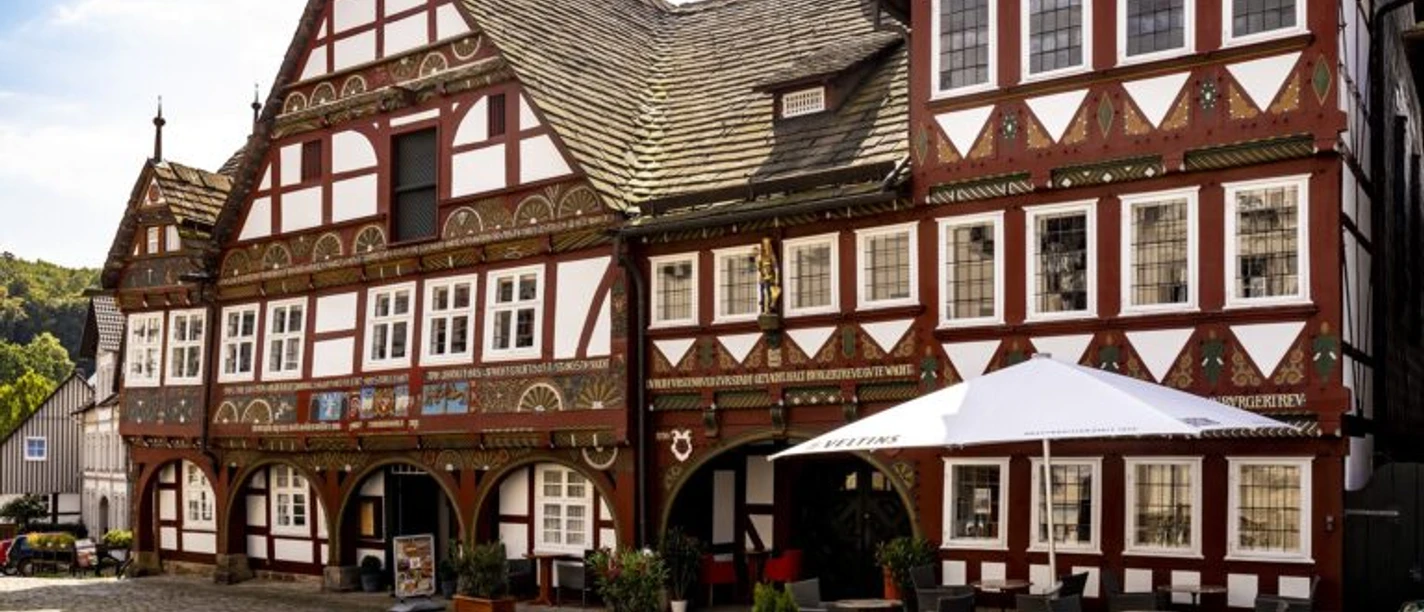 exterieurCL9A9992-2000-1-e1724078820263-768x502.jpg Historisches Fachwerkhaus mit verzierten Fassaden, Arkadengang und Außensitzbereich auf gepflastertem Platz.