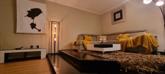 Wohnzimmer (2).jpg Ein gemütliches Wohnzimmer mit einem großen, beigen Sofa, gelben Decken und modernen Kunstwerken.