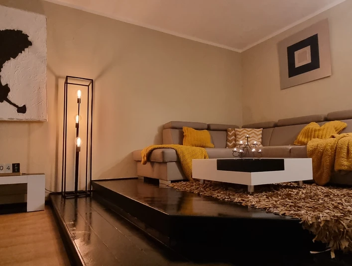 Wohnzimmer (2).jpg Ein gemütliches Wohnzimmer mit einem großen, beigen Sofa, gelben Decken und modernen Kunstwerken.