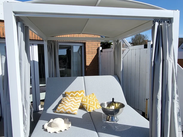 Daybed.jpg Ein modernes Daybed mit hellgrauen Polstern, umrahmt von weißen Vorhängen, ausgestattet für Entspannung.