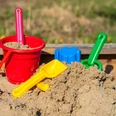 Ein Sandkasten voller Spielzeug mit einem roten Eimer, gelbem und grünem Spaten.