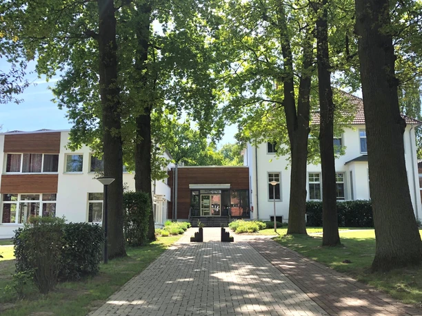 Denkhaus Loccum Zwei moderne Gebäude umgeben von hohen Bäumen, verbunden durch einen zentralen Eingangsbereich.