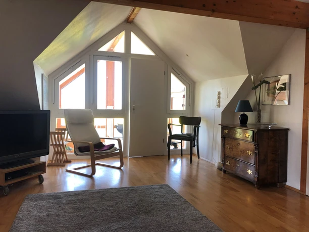 Bright attic room with wooden ceiling, large windows, TV, comfortable armchair and chest of drawers.