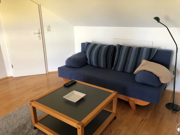 Bank met blauwe kussens, lichtgekleurd parket, salontafel met schaal, staande lamp in de woonkamer van een vakantiewoning.