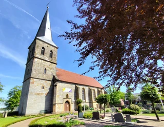 Ev.-ref.Kirche_Schapen©SG-Spelle.jpg Steinkirche mit hohem Turm und rotem Dach, umgeben von Friedhof und blühenden Bäumen im Sonnenschein