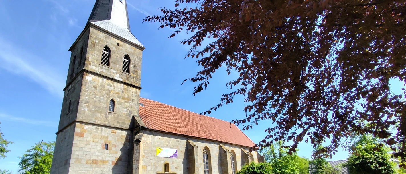 Ev.-ref.Kirche_Schapen©SG-Spelle.jpg Steinkirche mit hohem Turm und rotem Dach, umgeben von Friedhof und blühenden Bäumen im Sonnenschein