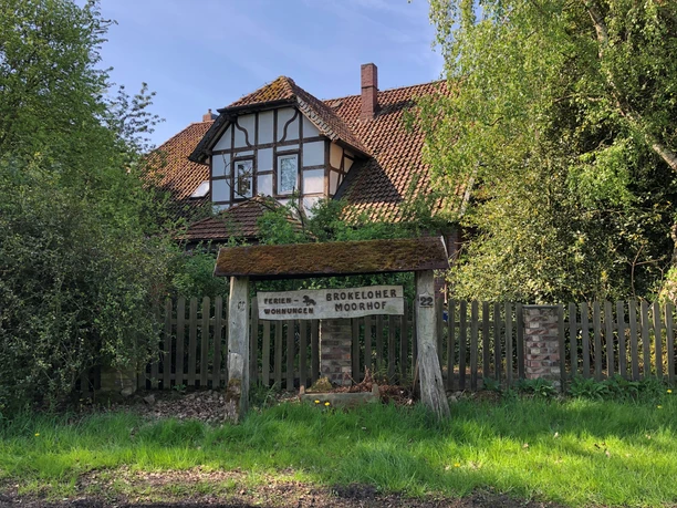 Half-timbered house with a red tiled roof, surrounded by green trees, with a rustic wooden sign in front of it.