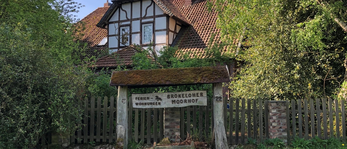 Brokeloher Moorhof Fachwerkhaus mit rotem Ziegeldach, umgeben von grünen Bäumen, mit einem rustikalen Holzschild davor.