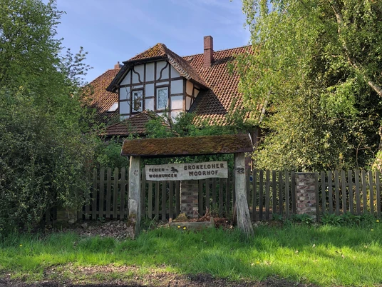 Brokeloher Moorhof Fachwerkhaus mit rotem Ziegeldach, umgeben von grünen Bäumen, mit einem rustikalen Holzschild davor.