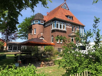 Pilgerherberge Altes Pfarrhaus Backsteinbau mit rotem Ziegeldach, weißen Gauben und Erkern in grüner Gartenumgebung.Brick building with red tiled roof, white dormers and bay windows in a green garden setting.Murstensbygning med rødt tegltag, hvide kviste og karnapper i et grønt havemiljø.Bakstenen gebouw met rood pannendak, witte dakkapellen en erkers in een groene tuin.