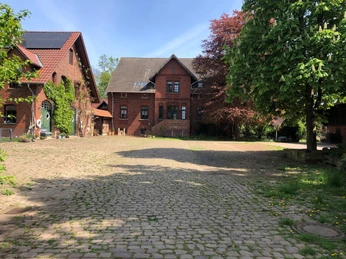 Hof Holthöfer Ein Bauernhof mit roten Backsteingebäuden und Kopfsteinpflaster umgeben von Bäumen an einem sonnigen Tag.