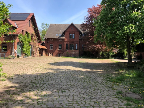 Hof Holthöfer Ein Bauernhof mit roten Backsteingebäuden und Kopfsteinpflaster umgeben von Bäumen an einem sonnigen Tag.