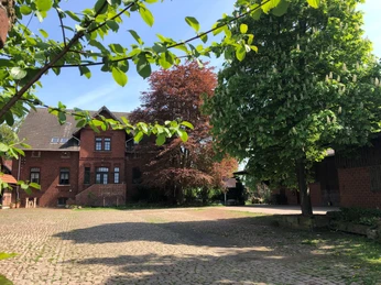 Hof Holthöfer Ein rotes Backsteingebäude auf einem Steinhof, umgeben von grünen und roten Laubbäumen.